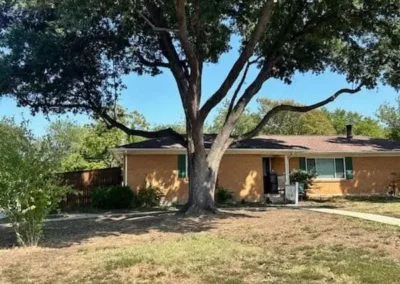 A large tree has been mostly cut down, with a worker using a chainsaw on the remaining stump and numerous logs and wood debris scattered around a house