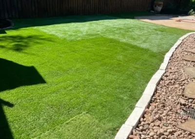 A lush green lawn, likely artificial turf, is immaculately laid out in a backyard, bordered by a decorative stone pathway and a dark wooden fence