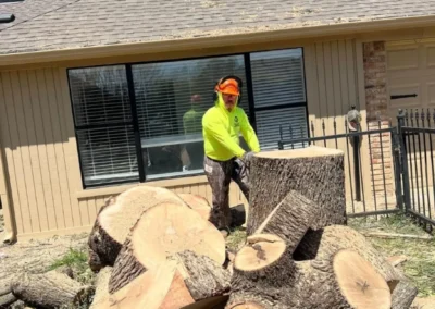 A man in a yellow shirt is cutting a tree outdoors, with a house and blue sky visible in the background