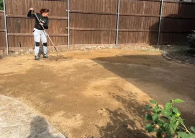 A person is raking dirt in a cleared backyard, preparing the ground for new landscaping, with a wooden fence and a house in the background