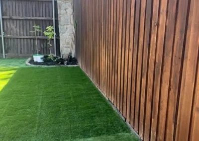 A view of a fenced yard featuring lush green artificial grass alongside a wooden fence and a small plant bed