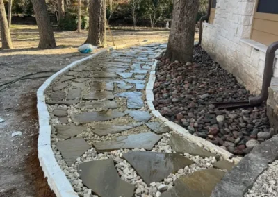 Curved stone pathway bordered by white concrete, lined with gravel and river rocks, under clear skies and bare trees