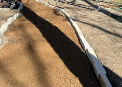 Two workers are engaged in laying down a path of gravel bordered by white stones in a sunny backyard, with a truck and trees in the background