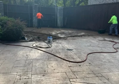 Two workers in bright shirts are in a backyard, one tending to a brown fence and the other working on the ground, with a large patio and hose in the foreground