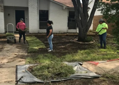 two are preparing the ground near a tree, and one is pushing a cart with sod rolls, in front of a white brick house on a sunny day